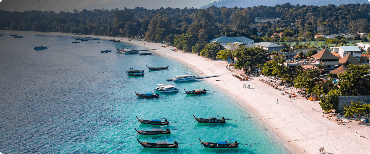 Boats anchored along the beach in Pattaya, Thailand with lush greenery and buildings nearby.