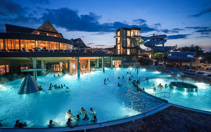 Tourists relaxing in Chochołow Thermal Baths with mountain view, evening lights, Zakopane transfers available.