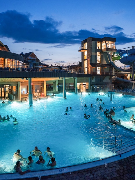 Tourists relaxing in Chochołow Thermal Baths with mountain view, evening lights, Zakopane transfers available.