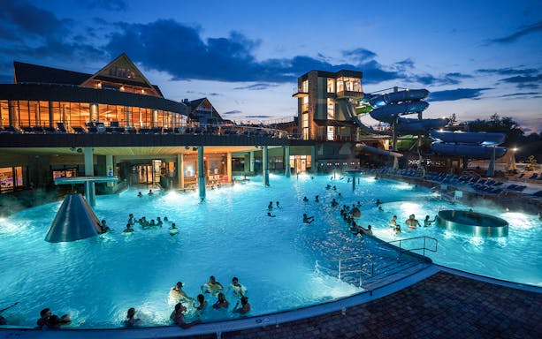 Tourists relaxing in Chochołow Thermal Baths with mountain view, evening lights, Zakopane transfers available.