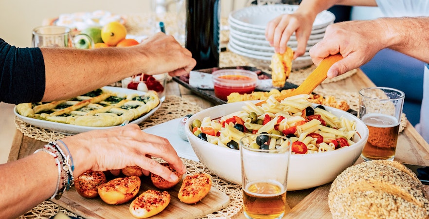 Pasta salad and bread on a dining table with people serving food.