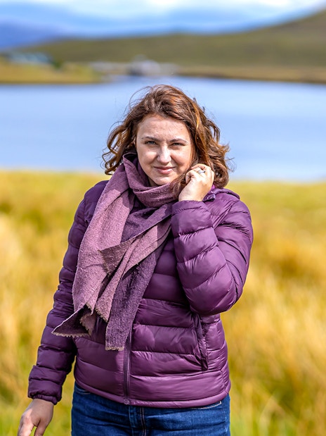 Person standing by a lake in the Isle of Skye, surrounded by grassy hills.