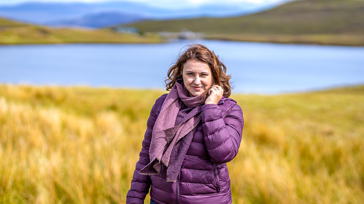 Person standing by a lake in the Isle of Skye, surrounded by grassy hills.