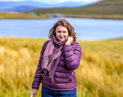 Person standing by a lake in the Isle of Skye, surrounded by grassy hills.