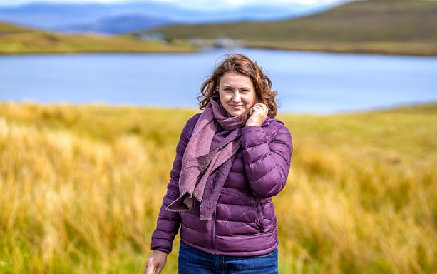 Person standing by a lake in the Isle of Skye, surrounded by grassy hills.