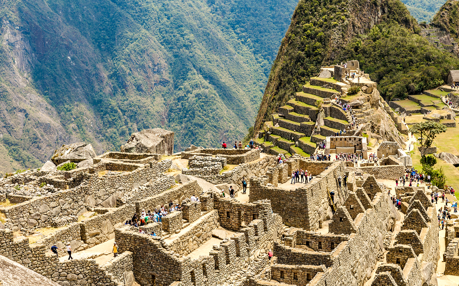 Tourists exploring the Sacred Plaza at Machu Picchu, Peru, with terraced mountains in the background.