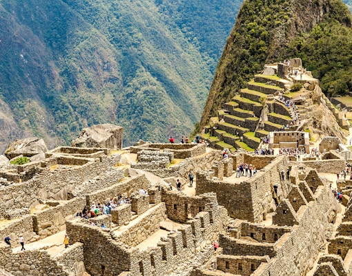 Tourists exploring the Sacred Plaza at Machu Picchu, Peru, with terraced mountains in the background.