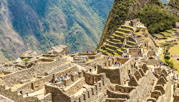 Tourists exploring the Sacred Plaza at Machu Picchu, Peru, with terraced mountains in the background.