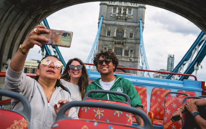 Tourists taking a selfie on a London hop-on hop-off bus near Tower Bridge.