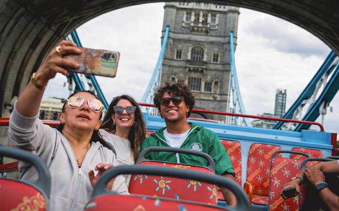 Tourists taking a selfie on a London hop-on hop-off bus near Tower Bridge.