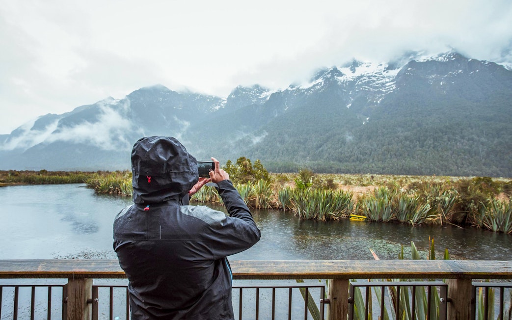 Person photographing misty mountains and lake in Milford Sound, New Zealand.