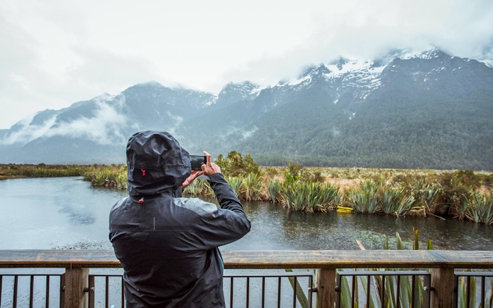 Person photographing misty mountains and lake in Milford Sound, New Zealand.