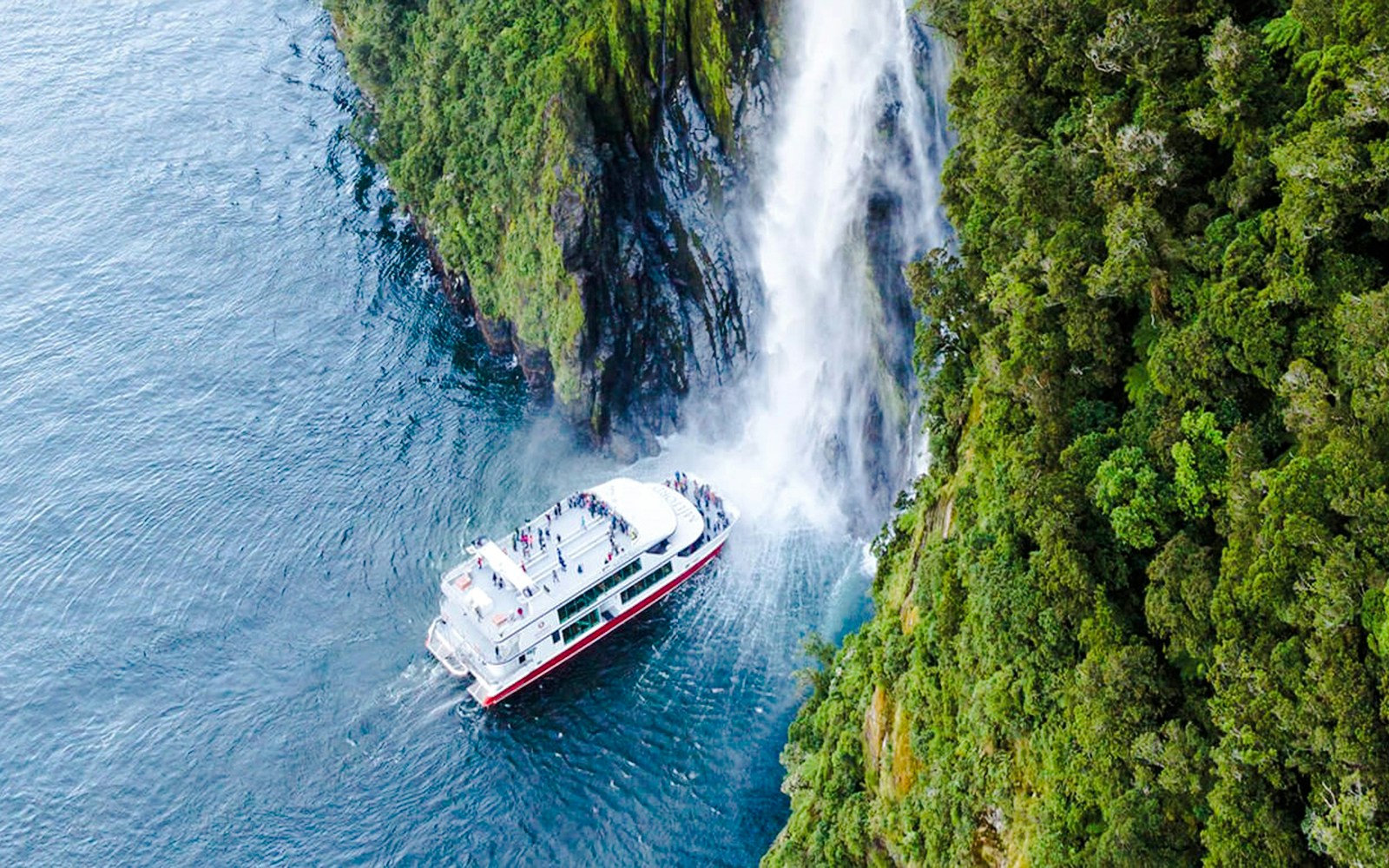 Cruise boat near waterfall in Milford Sound's lush landscape.