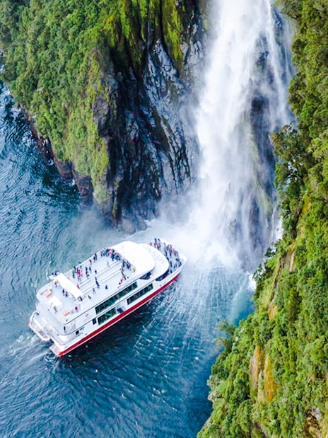 Cruise boat near waterfall in Milford Sound's lush landscape.