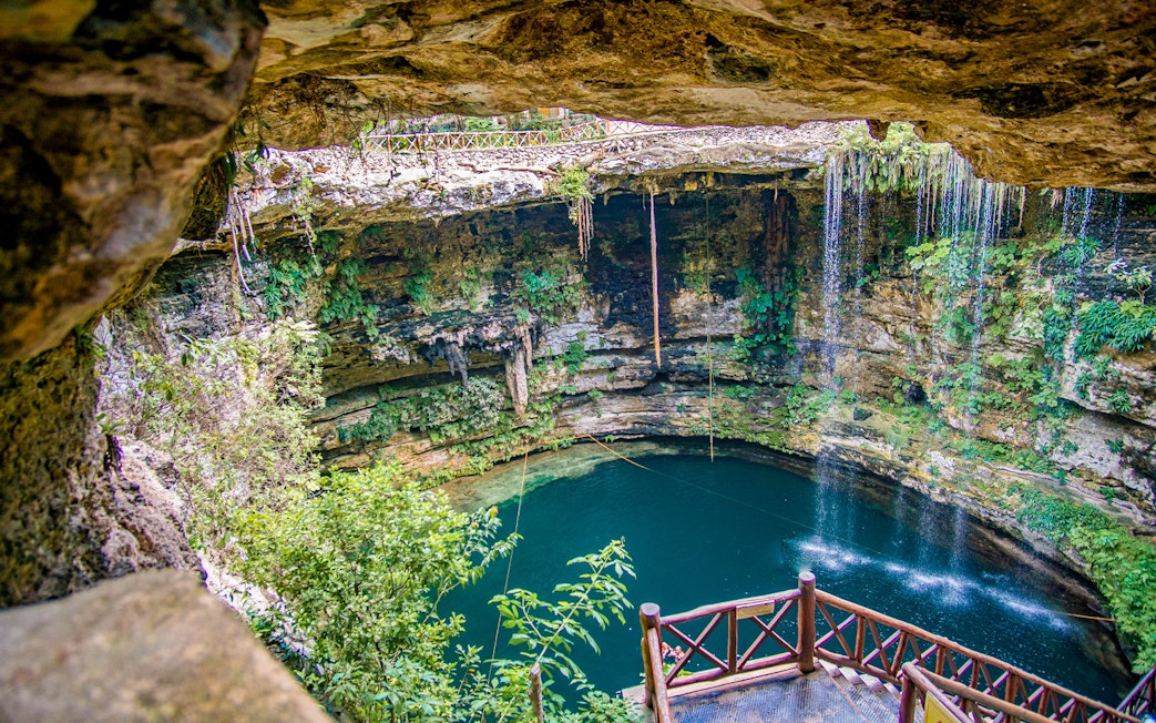 Entrance to a cenote with wooden railing, surrounded by lush vegetation and rock formations.
