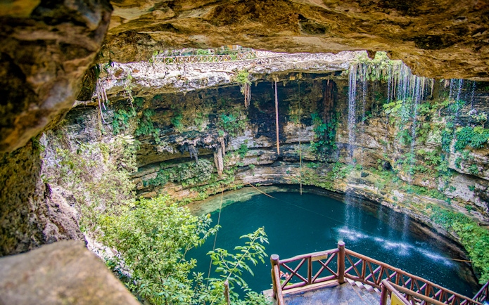 Entrance to a cenote with wooden railing, surrounded by lush vegetation and rock formations.