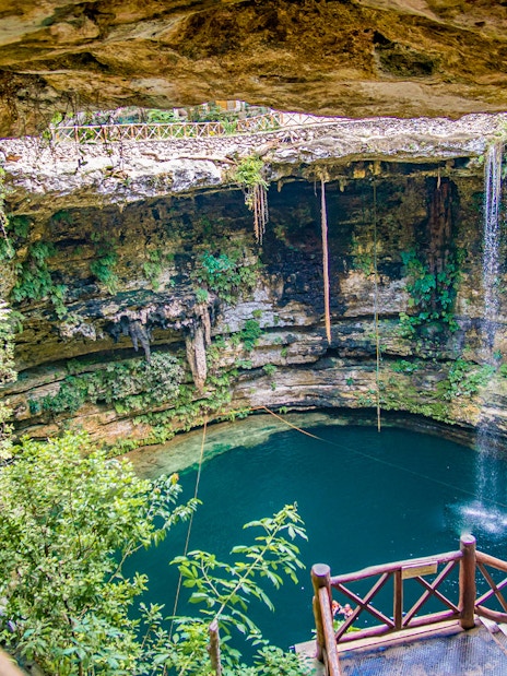 Entrance to a cenote with wooden railing, surrounded by lush vegetation and rock formations.