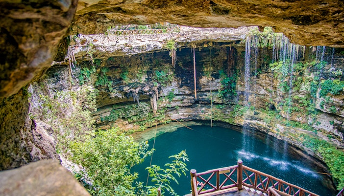 Entrance to a cenote with wooden railing, surrounded by lush vegetation and rock formations.
