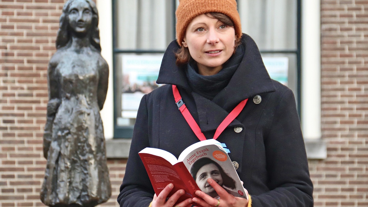 Tour guide holding a book near Anne Frank statue in Amsterdam.