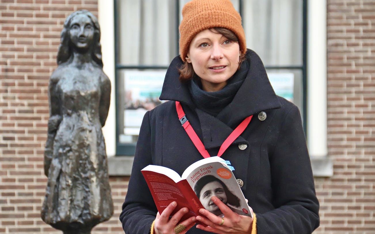 Tour guide holding a book near Anne Frank statue in Amsterdam.