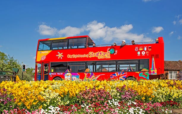 Red double-decker bus on City Sightseeing tour in Stratford-upon-Avon, passing colorful flower beds.