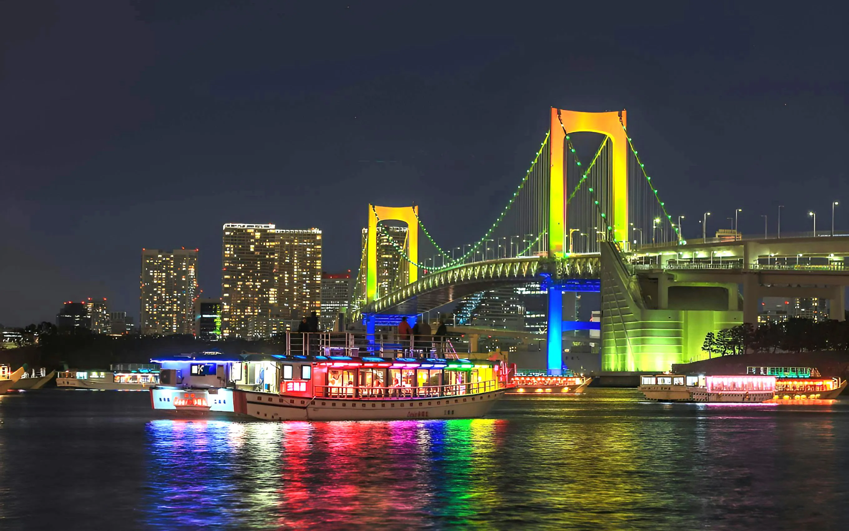Rainbow Bridge in Tokyo illuminated at night with colorful boats on the water.