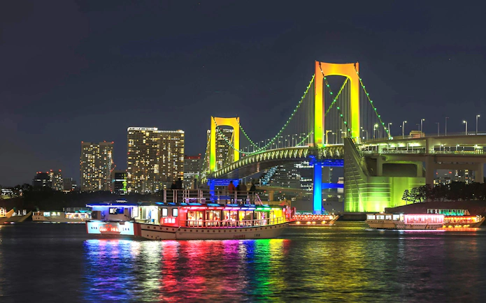Rainbow Bridge in Tokyo illuminated at night with colorful boats on the water.