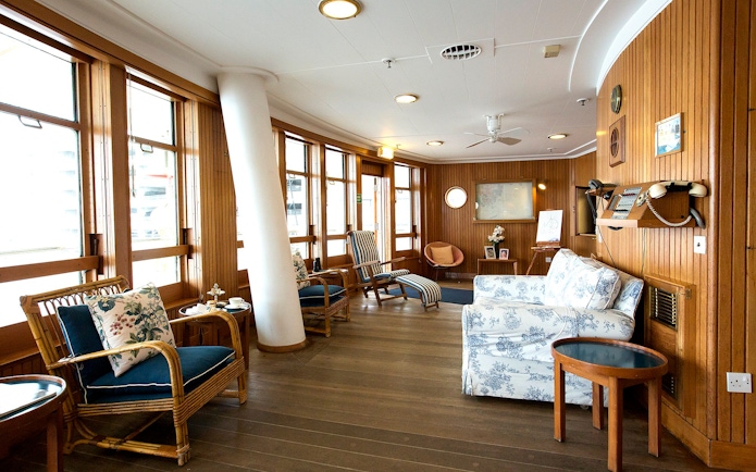 Wood-paneled sun lounge with wicker chairs and floral cushions on a cruise ship.