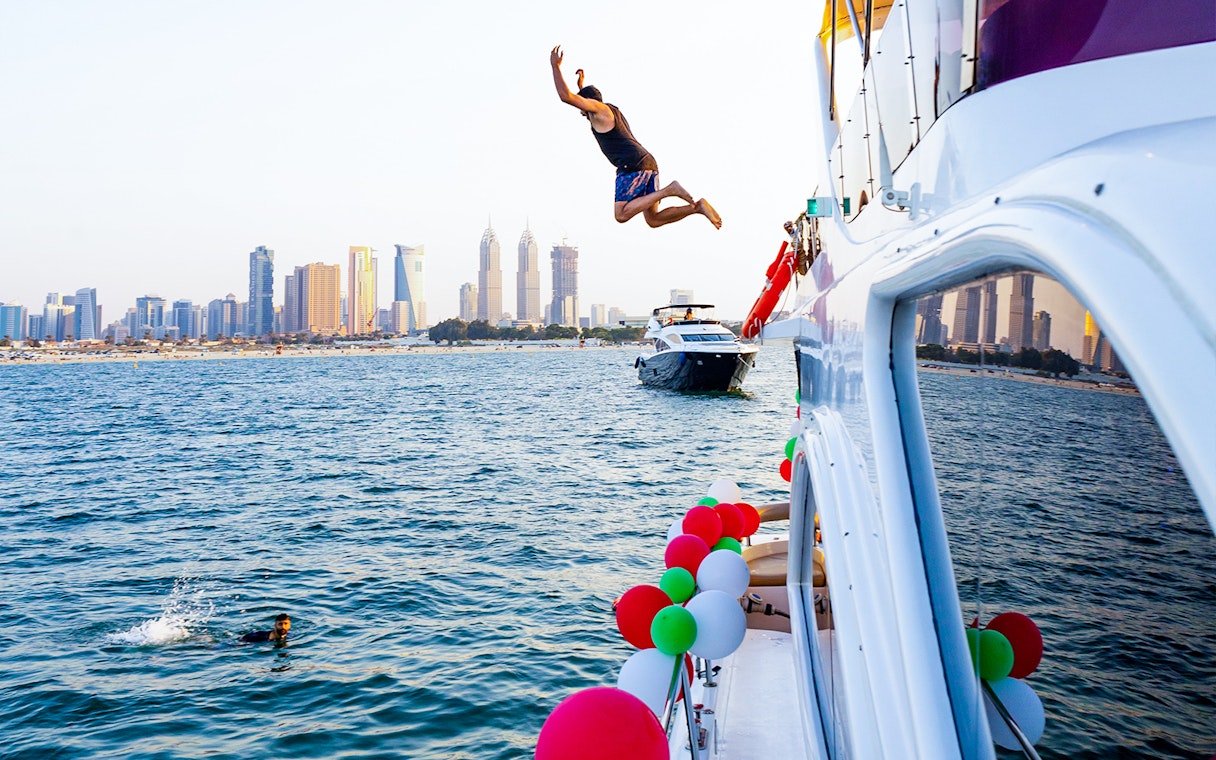 Man jumping off yacht into water with Dubai skyline in background.