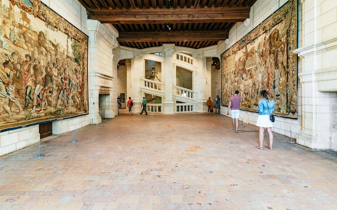 Visitors near the helix staircase in Chambord Castle, France, with tapestries on the walls.
