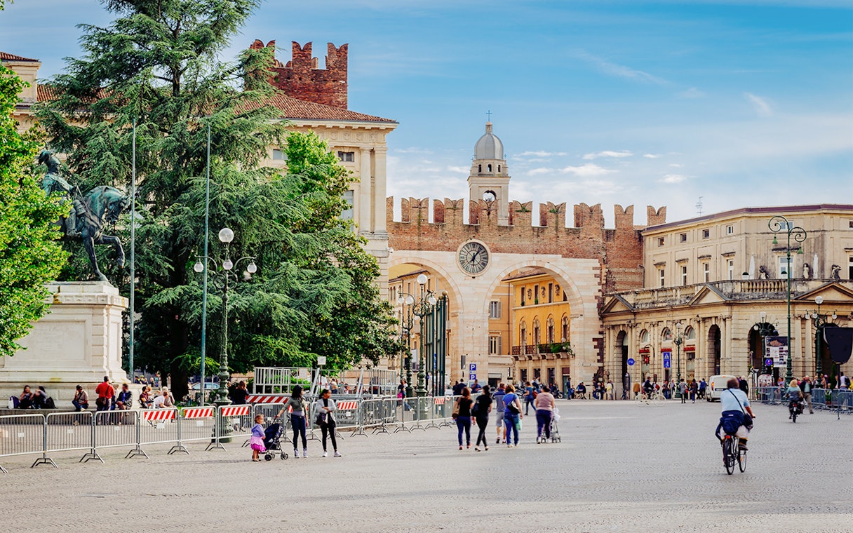 Medieval gates and clock tower on Piazza Bra, Verona, Italy, with people walking nearby.