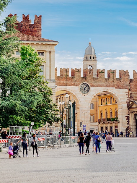 Medieval gates and clock tower on Piazza Bra, Verona, Italy, with people walking nearby.