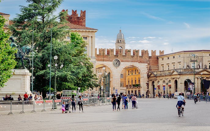 Medieval gates and clock tower on Piazza Bra, Verona, Italy, with people walking nearby.