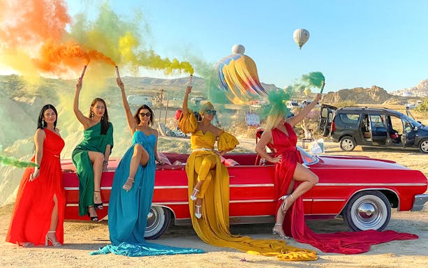 Women in colorful dresses with smoke sticks on a red vintage car in Cappadocia, hot air balloons in background.