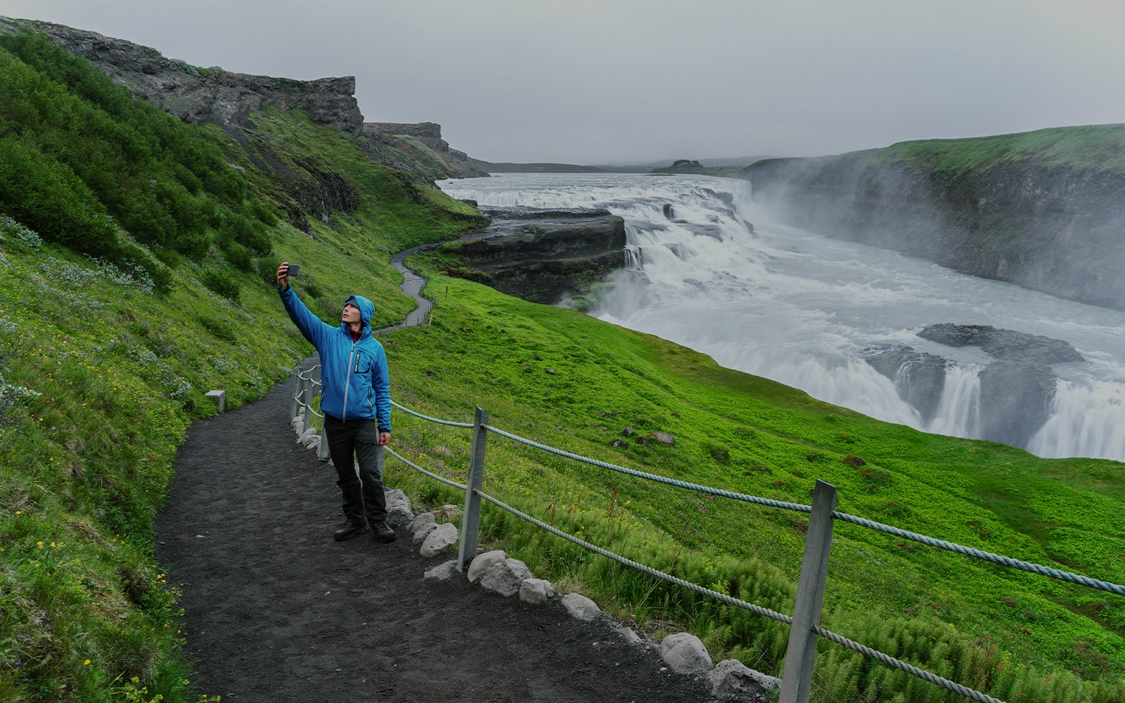 Person taking a selfie near Gullfoss Falls, Iceland, with lush green landscape.