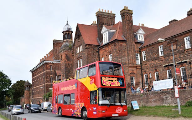 Red double-decker sightseeing bus in front of historic brick building in Norwich.