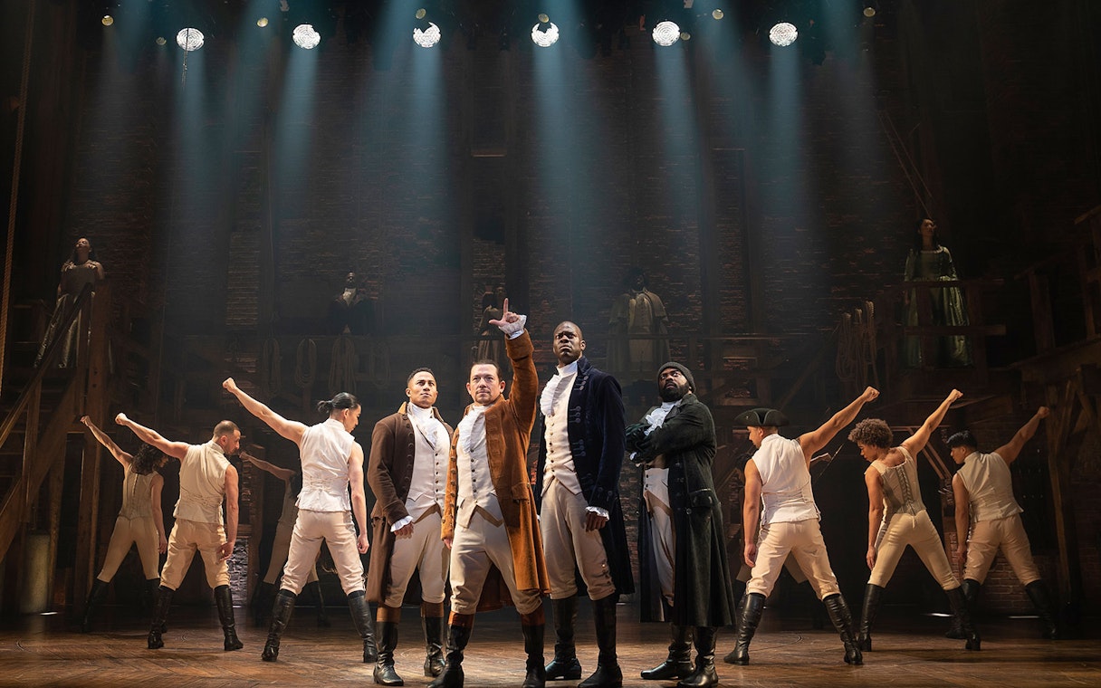 Stage performance of Hamilton with actors in period costumes under dramatic lighting.