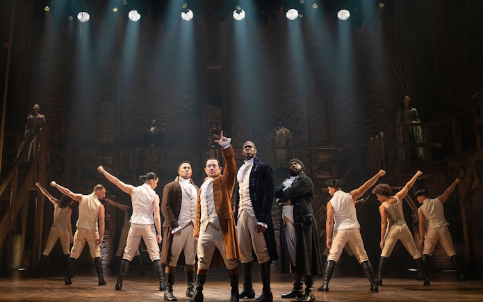 Stage performance of Hamilton with actors in period costumes under dramatic lighting.