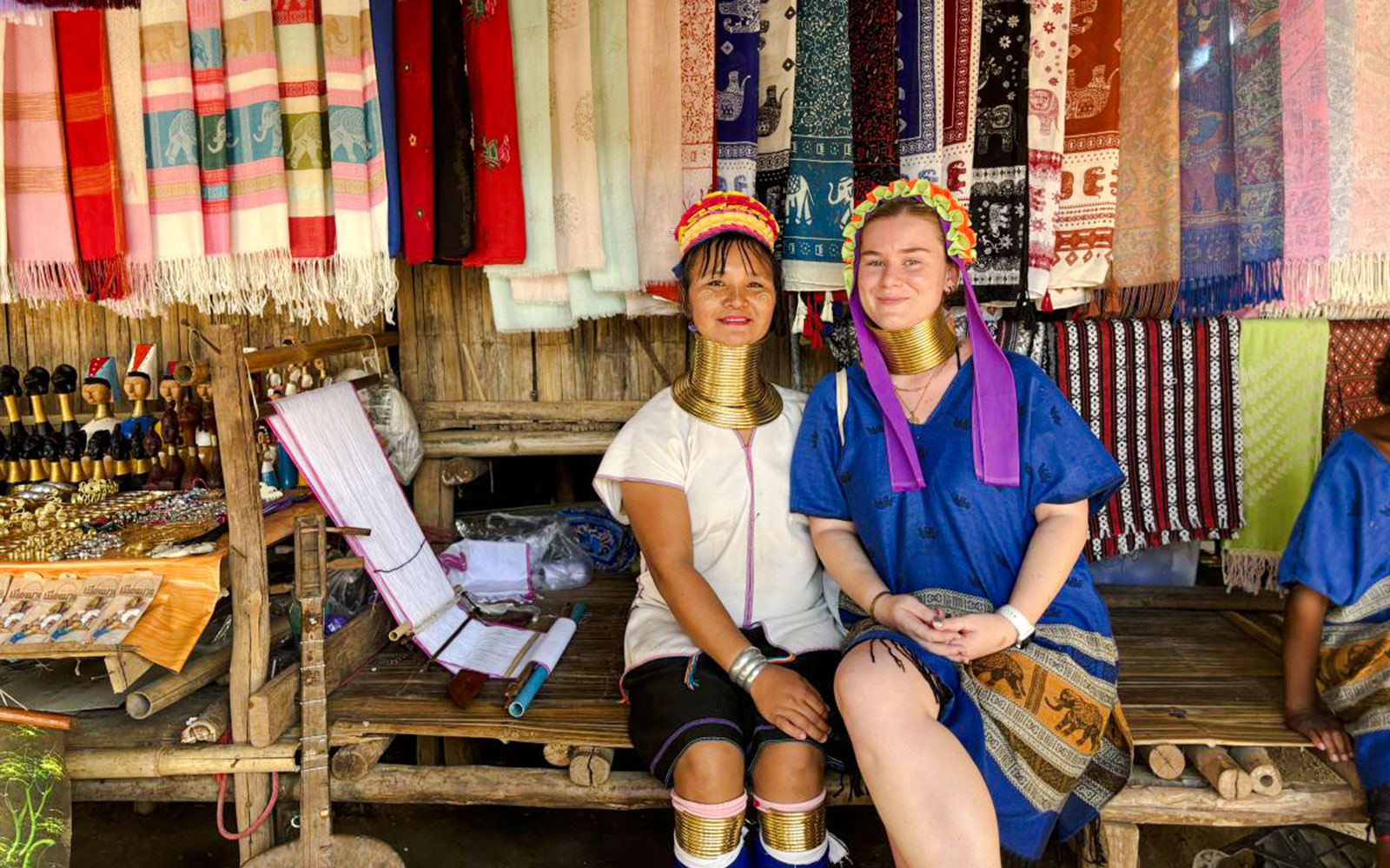 Women in traditional attire with neck rings at a Chiang Mai market stall displaying colorful textiles.