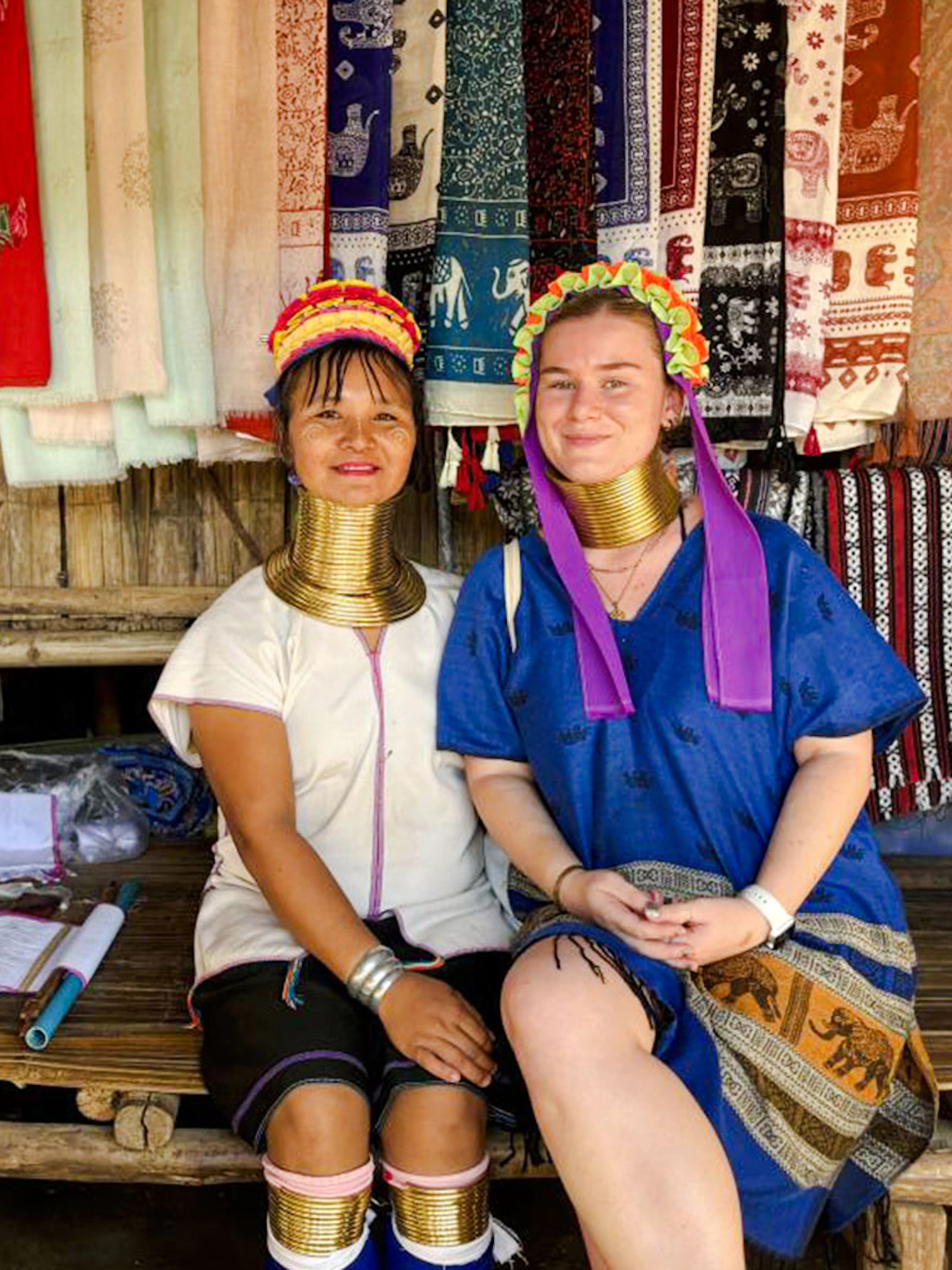 Women in traditional attire with neck rings at a Chiang Mai market stall displaying colorful textiles.