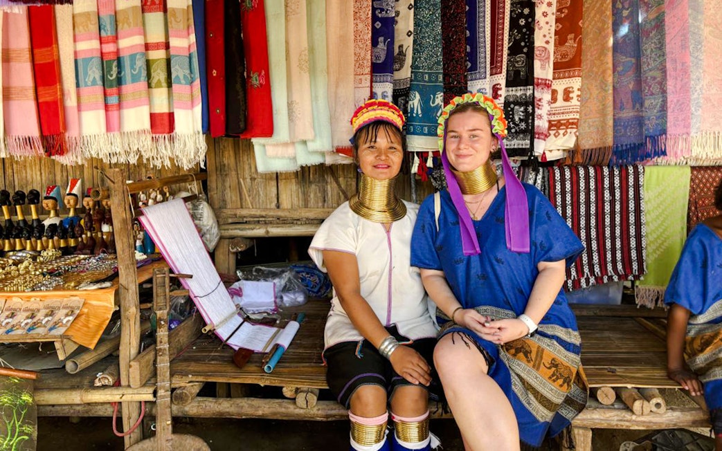 Women in traditional attire with neck rings at a Chiang Mai market stall displaying colorful textiles.