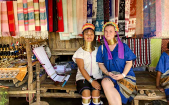 Women in traditional attire with neck rings at a Chiang Mai market stall displaying colorful textiles.