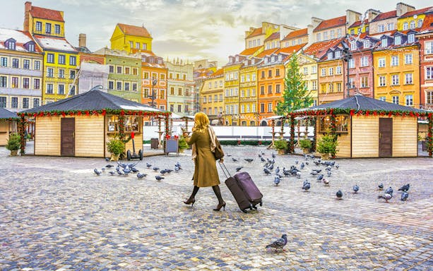 Woman with suitcase walking through Warsaw's Old Town square with colorful buildings.