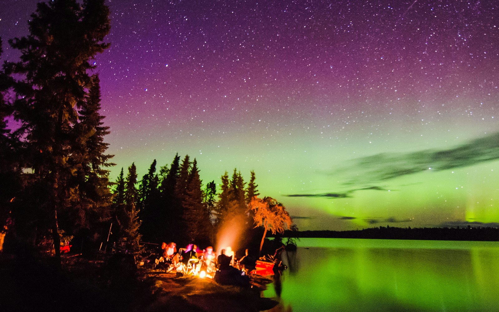 Campfire by a lake under the Northern Lights with silhouetted trees.