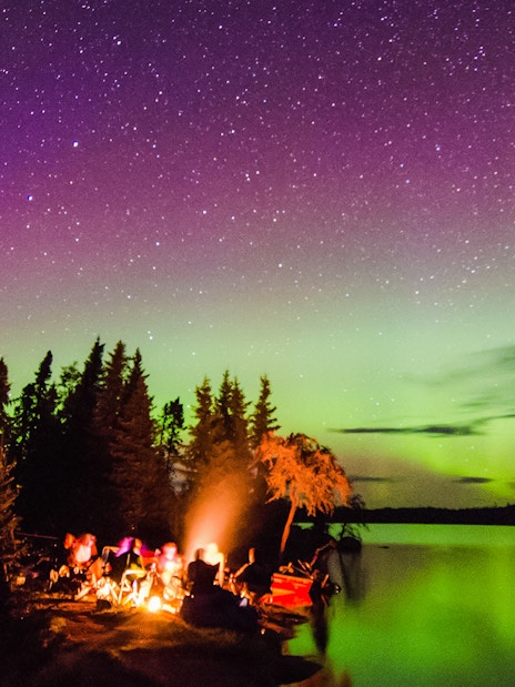 Campfire by a lake under the Northern Lights with silhouetted trees.