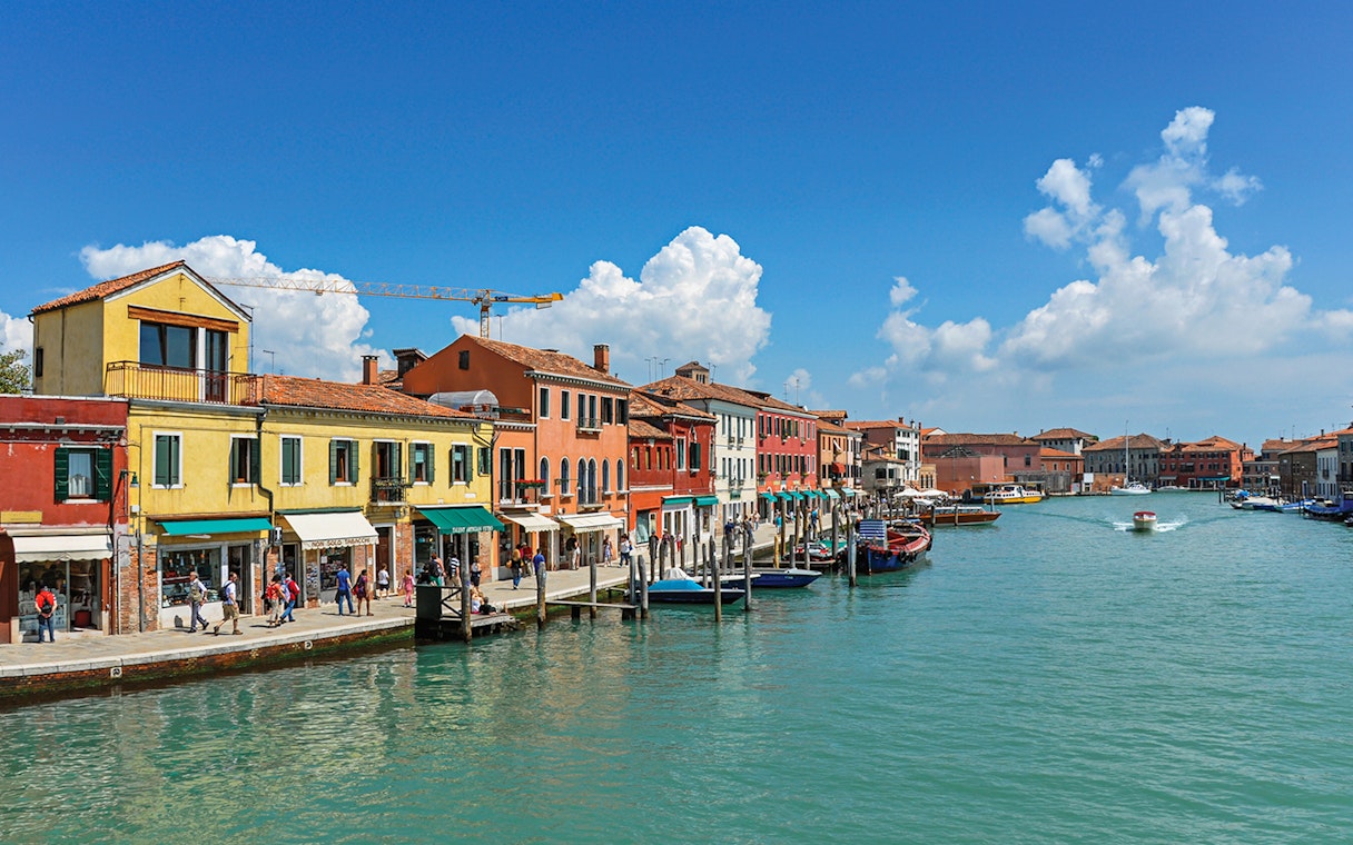 Colorful buildings along a canal in Murano, Italy, with boats and tourists.
