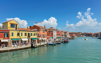 Colorful buildings along a canal in Murano, Italy, with boats and tourists.