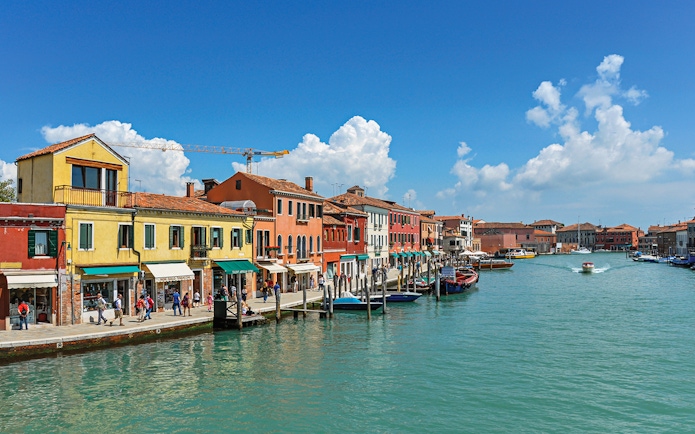 Colorful buildings along a canal in Murano, Italy, with boats and tourists.