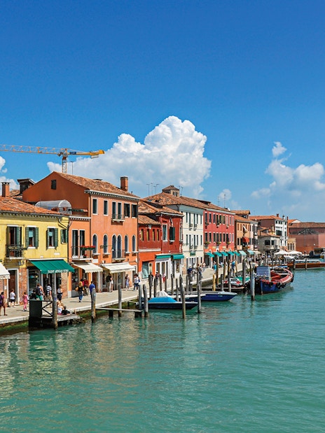 Colorful buildings along a canal in Murano, Italy, with boats and tourists.