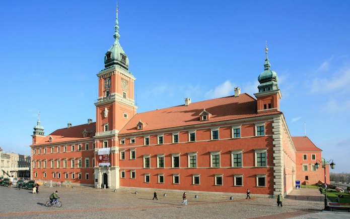 Royal Castle in Castle Square, Warsaw, Poland, with people walking and cycling nearby.
